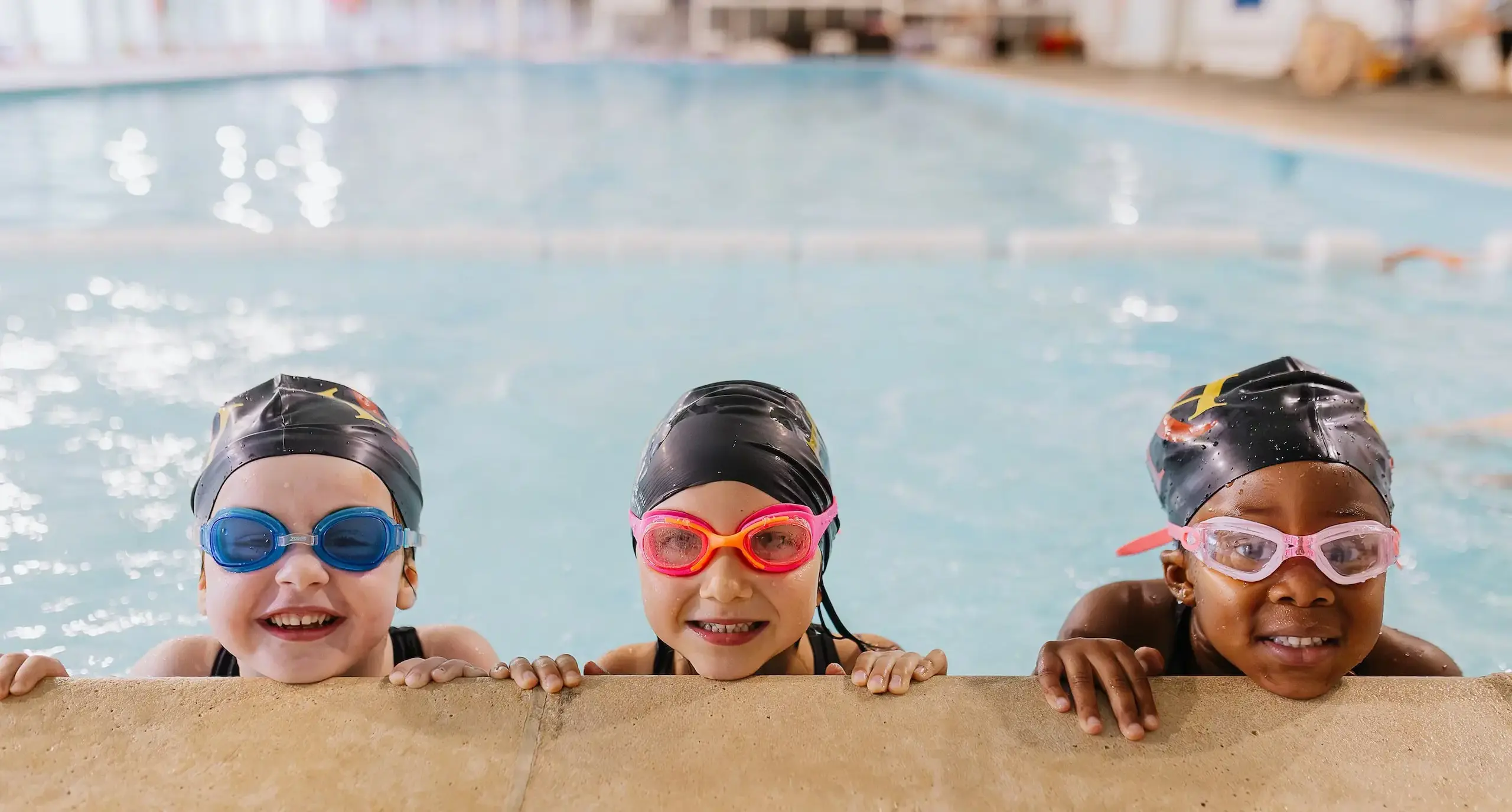 Three students peeking up from a swimming pool, all wearing goggles and swimming caps